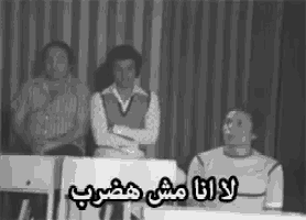 a black and white photo of three men sitting at a table with arabic writing on it .