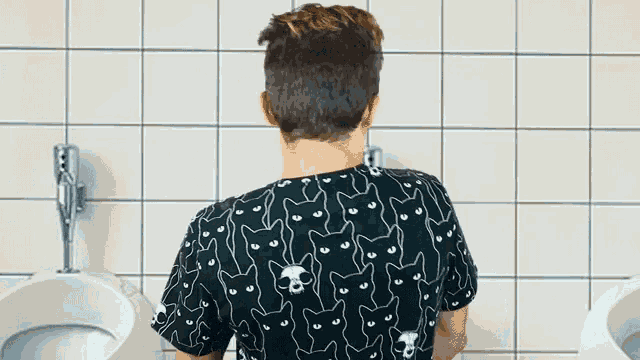 a man in a black shirt with cats on it is urinating in a bathroom
