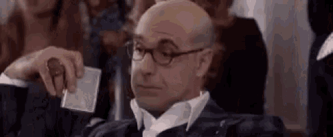 a bald man wearing glasses and a suit is sitting in a chair holding a piece of paper .