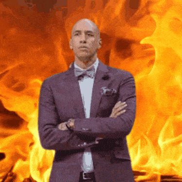 a man in a suit and bow tie stands with his arms crossed in front of flames