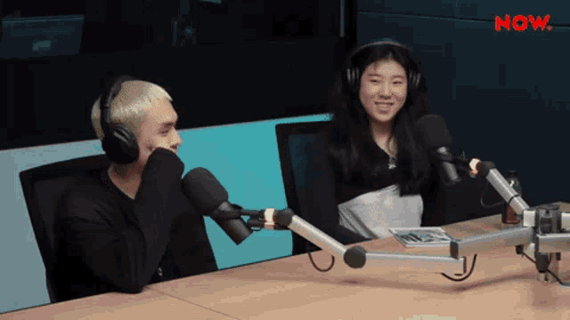 a man and a woman are sitting at a table with microphones and the word now on the wall