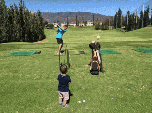 a little boy watches a man swing a golf club at a golf ball