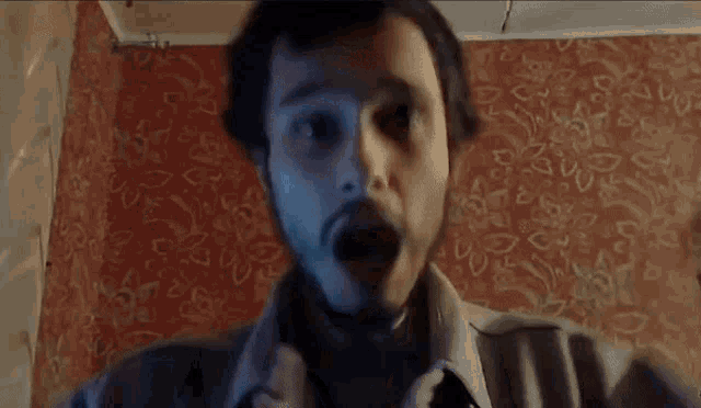 a man with a beard is sitting in front of a wall with a patterned wallpaper .