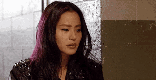 a woman with purple hair is standing next to a brick wall and crying .