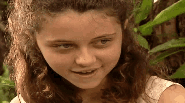 a young girl with curly hair and a white shirt is smiling and looking at the camera .