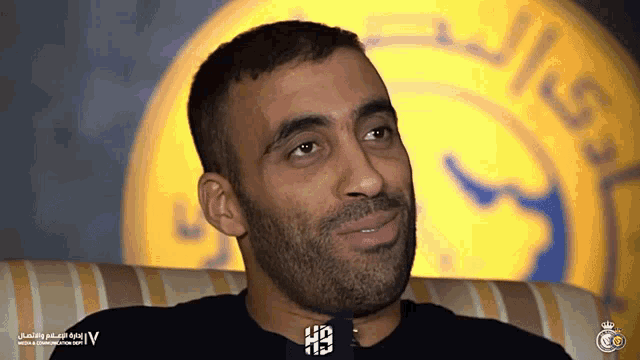 a man with a beard is sitting in front of a yellow and blue logo that says ' saudi arabia ' on it