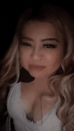 a woman with long blonde hair and a nose ring is wearing a white tank top and looking at the camera .