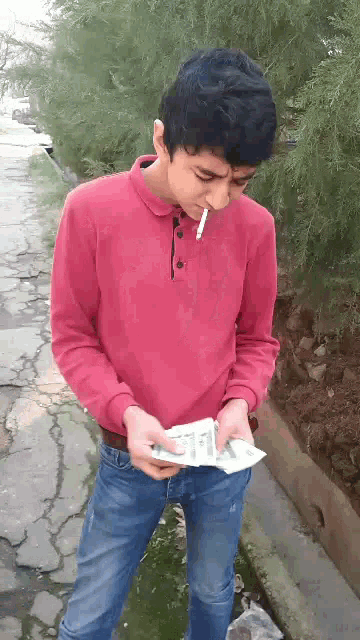 a man in a red shirt is smoking a cigarette while holding a stack of money .