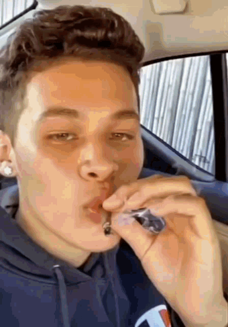 a young man is smoking a cigarette in a car while wearing a blue hoodie .