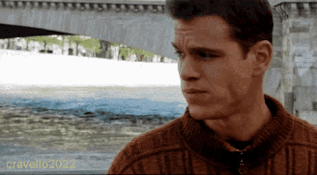 a man wearing a brown sweater is standing in front of a body of water