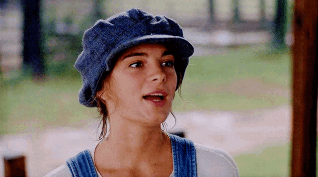 a woman wearing a blue hat and overalls is making a funny face
