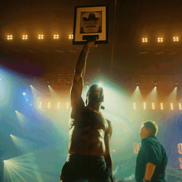 a shirtless man holds up a framed picture of a man named michael jackson in a dark room
