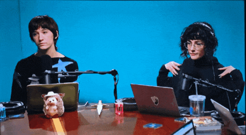 two people are sitting at a table with laptops and microphones in front of a blue wall