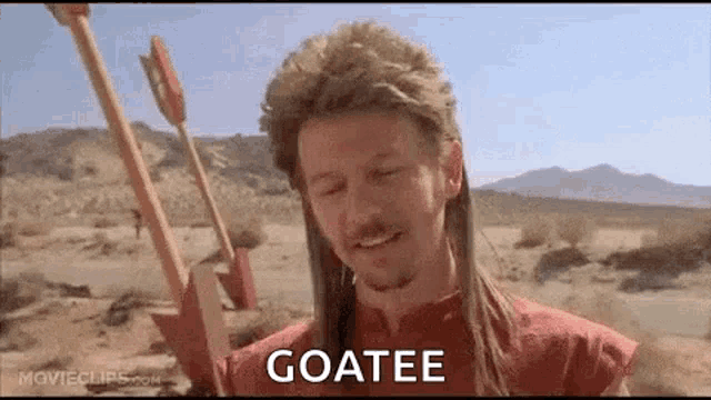 a man in a mullet is holding an arrow in the desert and says `` goatee '' .