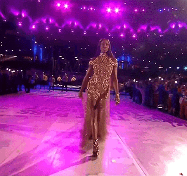a woman in a gold dress walks on a stage with purple lights
