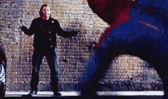 a man in a black jacket is standing next to a brick wall while a spiderman is standing behind him .