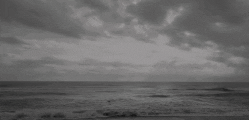 a black and white photo of a beach with a cloudy sky and waves crashing on the shore .