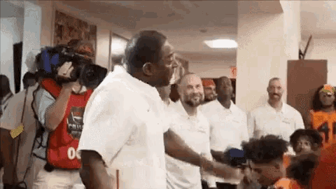 a group of men are shaking hands in a hallway while a cameraman takes a picture of them .