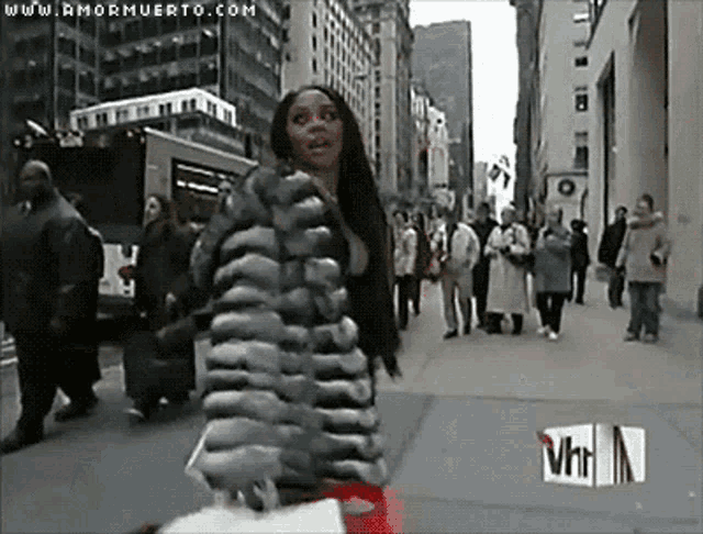 a woman in a fur coat is walking down a city street with a vh1 logo in the background
