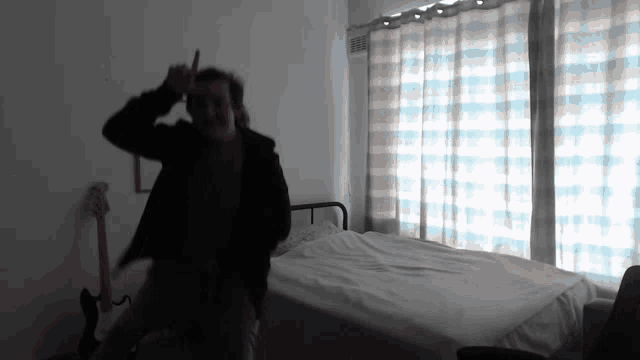 a man in a black jacket stands in front of a bed with a guitar hanging on the wall