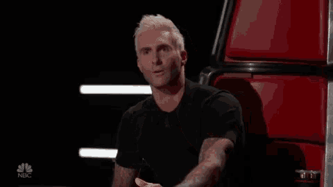 a man with white hair is sitting in front of a red chair with his hands folded in front of his face .