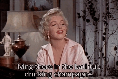 a woman in a pink shirt is laying in a bathtub drinking champagne