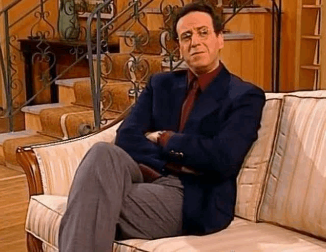 a man in a suit and tie is sitting on a couch with his arms crossed and looking at the camera .