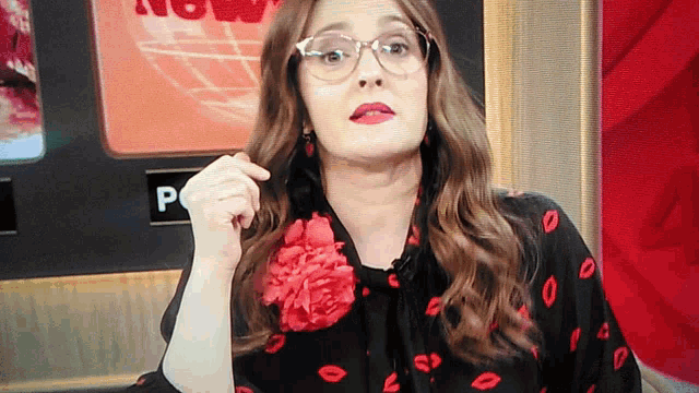a woman wearing glasses and a red flower in her hair stands in front of a screen that says now