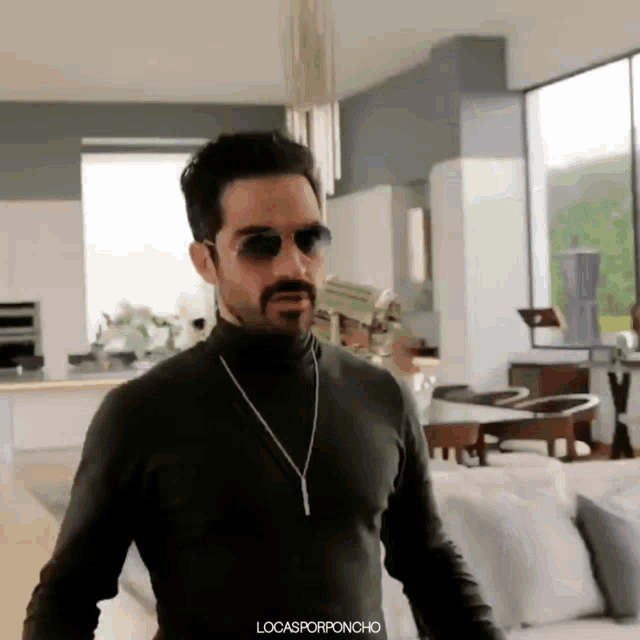 a man with a beard wearing sunglasses and a turtleneck is standing in a living room .