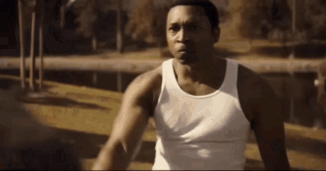 a man in a white tank top is standing in a park with trees in the background .