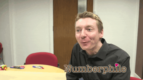 a man sitting at a table with the name numberphile written on his shirt