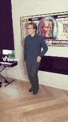 a man in a blue shirt is standing in a living room in front of a poster of a dollar bill .
