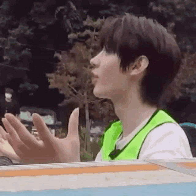 a young man wearing a green vest and a white shirt is sitting on a bench with his hands outstretched .