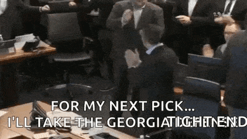 a man in a suit is sitting at a table in front of a crowd of people and talking to them .