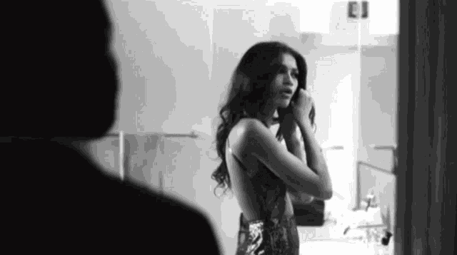 a black and white photo of a woman in a dress looking at herself in a mirror .