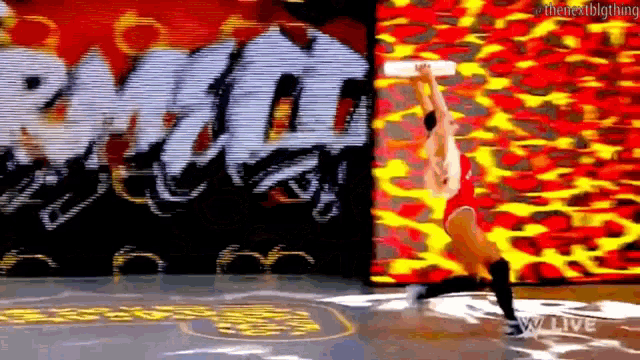 a wrestler is jumping in the air while holding a trophy in front of a wall with graffiti on it .