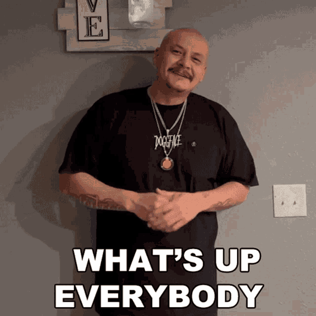 a man wearing a dogface shirt is standing in front of a wall with the words what 's up everybody written on it