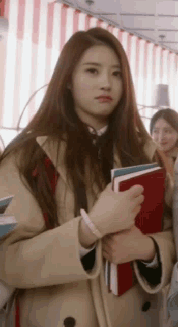 a girl in a trench coat is holding a stack of books in her hands