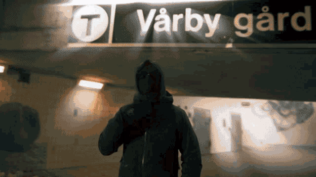 a man in a hooded jacket stands in front of a varby gard sign