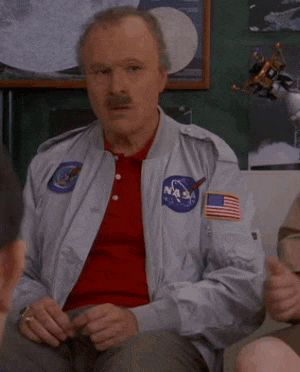 a man wearing a nasa jacket and a red shirt is sitting in a room with other people .