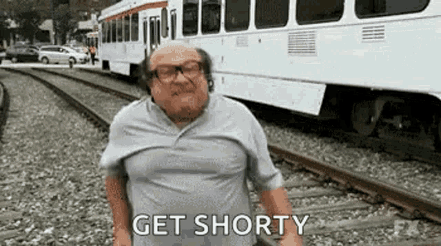 a man is standing on train tracks in front of a train and saying `` get shorty '' .