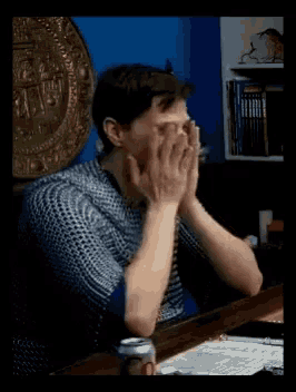 a man covering his face with his hands while sitting at a desk with a can of beer in front of him