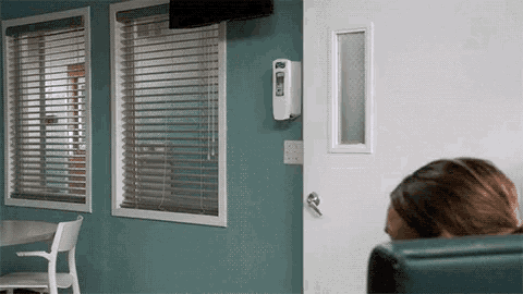 a woman sits in a chair in front of a window with blinds and a hand sanitizer dispenser