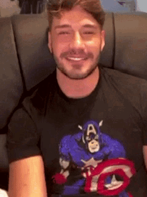 a man is sitting on a couch wearing a captain america t-shirt and smiling .