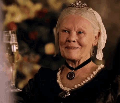 an older woman wearing a tiara and a choker holds a glass of champagne