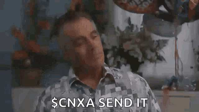 a man in a plaid shirt is sitting in front of balloons and flowers and says `` scnxa send it ''