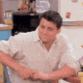 a man in a white shirt is sitting at a table in a kitchen .