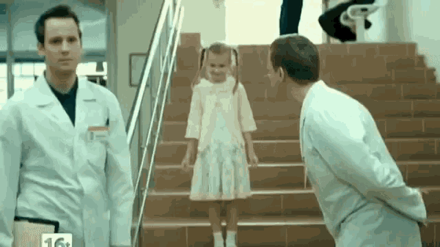 a little girl in a pink dress is standing on a set of stairs next to two men in lab coats .
