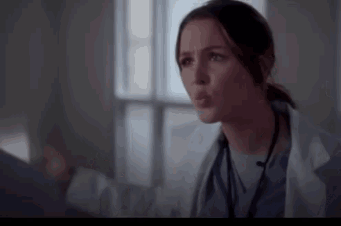a woman in a lab coat and scrubs is standing in front of a window in a room .