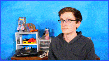 a young man wearing glasses stands in front of a blue wall and a shelf with a picture of two men on it